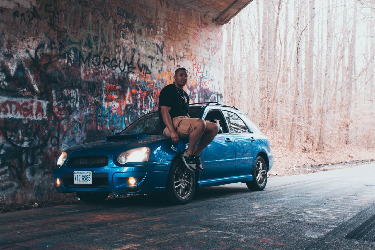 person sitting on a blue car under a graffiti covered bridge in willia