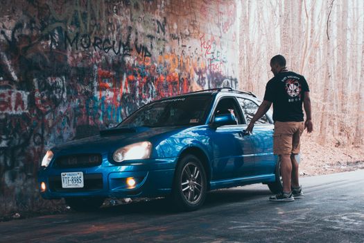 A man stands beside a blue car in a graffiti-covered tunnel in Williamsburg, VA.