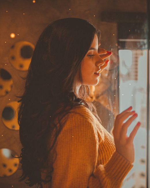 Side profile of a woman in knitwear, standing by a window, looking contemplative.