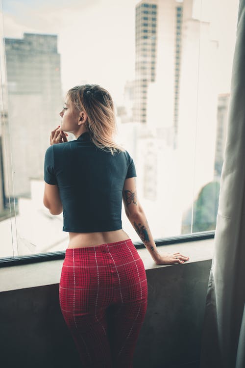 Back View Photo of Woman In Black Top Standing Beside Window Looking Outside