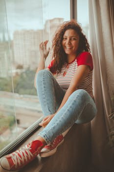 一位卷发微笑的女人，穿着牛仔裤和条纹衬衫，在窗前摆着姿势，散发着幸福感