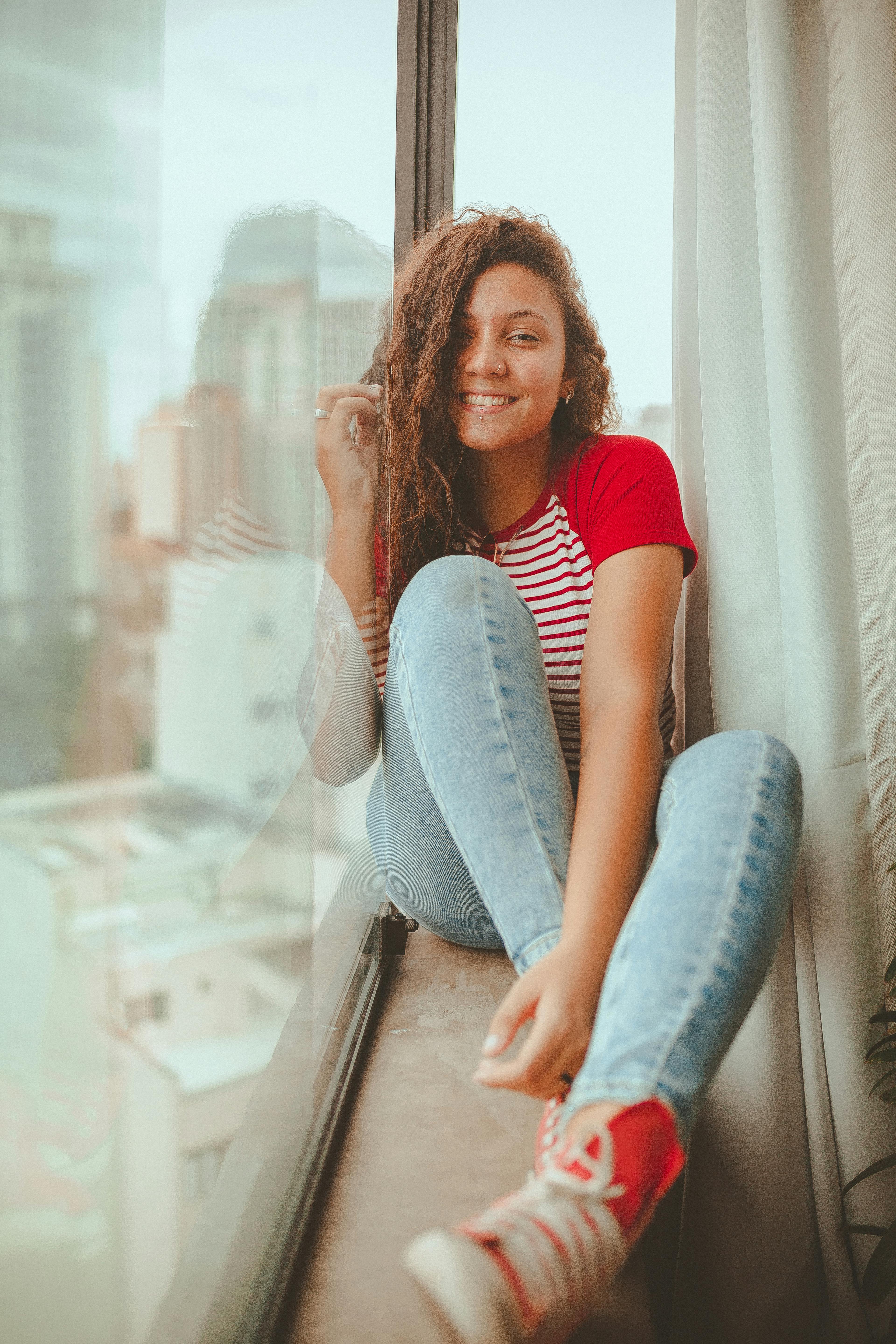 一位卷发的年轻女子穿着休闲装，坐在窗前微笑
