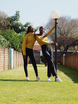Two stylish young women pose outdoors in a garden setting, showcasing modern fashion in bright sunlight.
