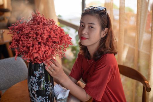 Asian woman in casual attire sitting indoors with flowers, looking at camera, smiling.