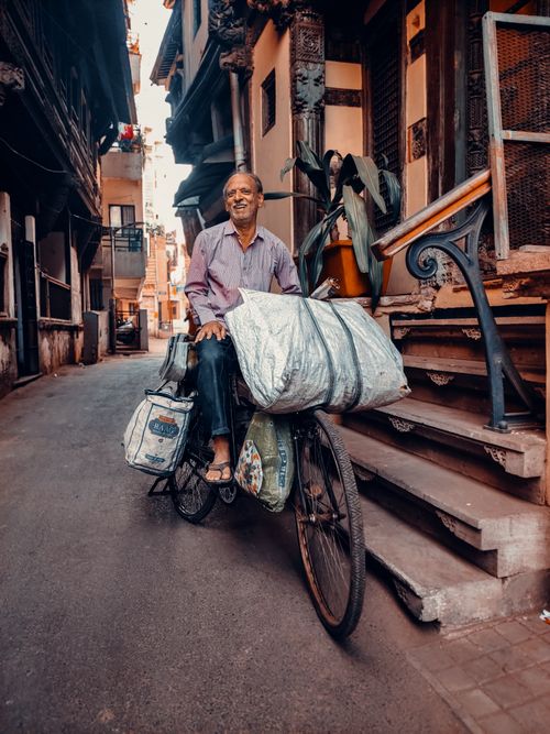 Free Elderly man with bags on a bicycle, smiling on a narrow street in Ahmedabad, India. Stock Photo