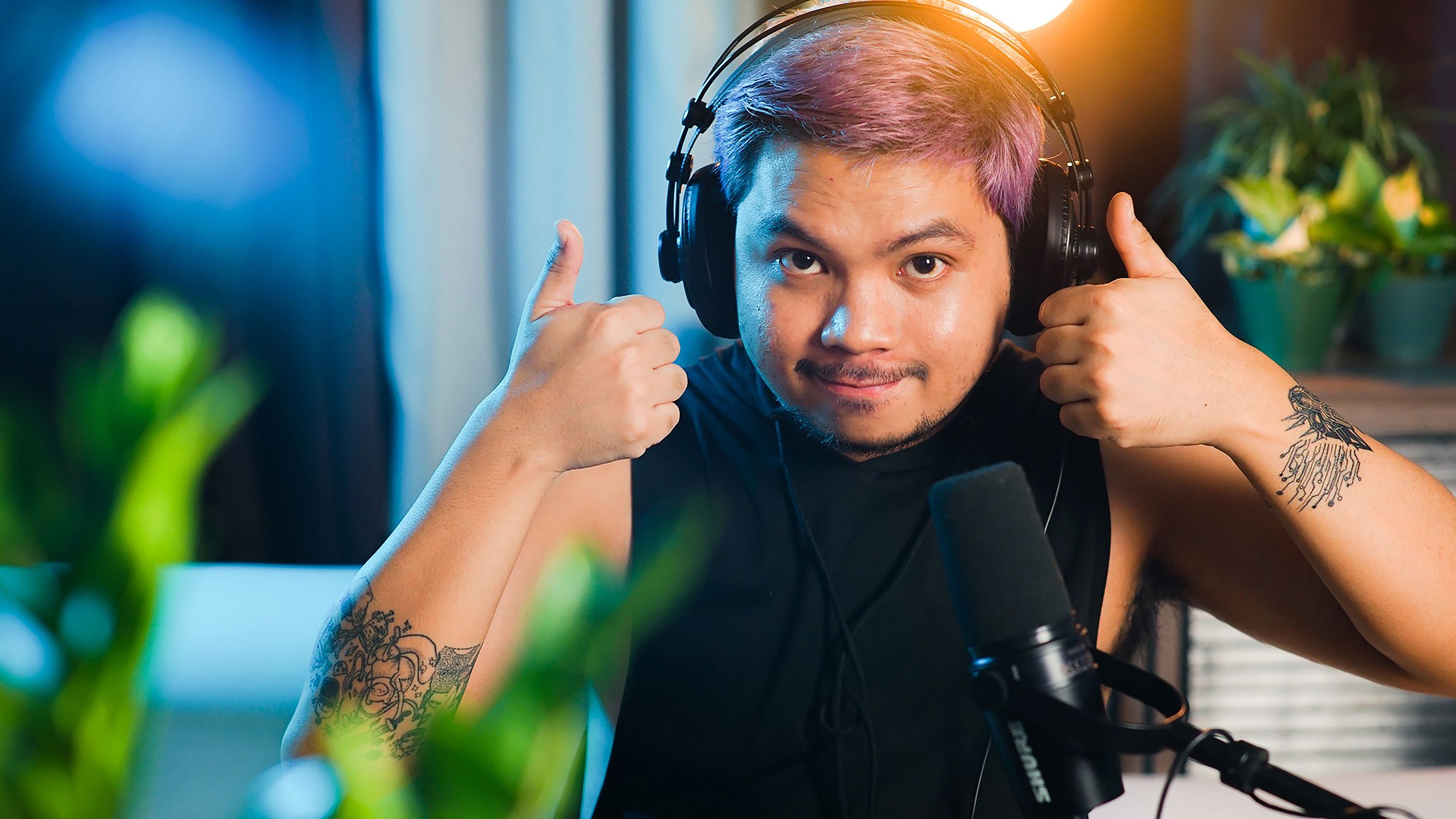 A man in a black tank top records a podcast in a studio while giving a thumbs up.