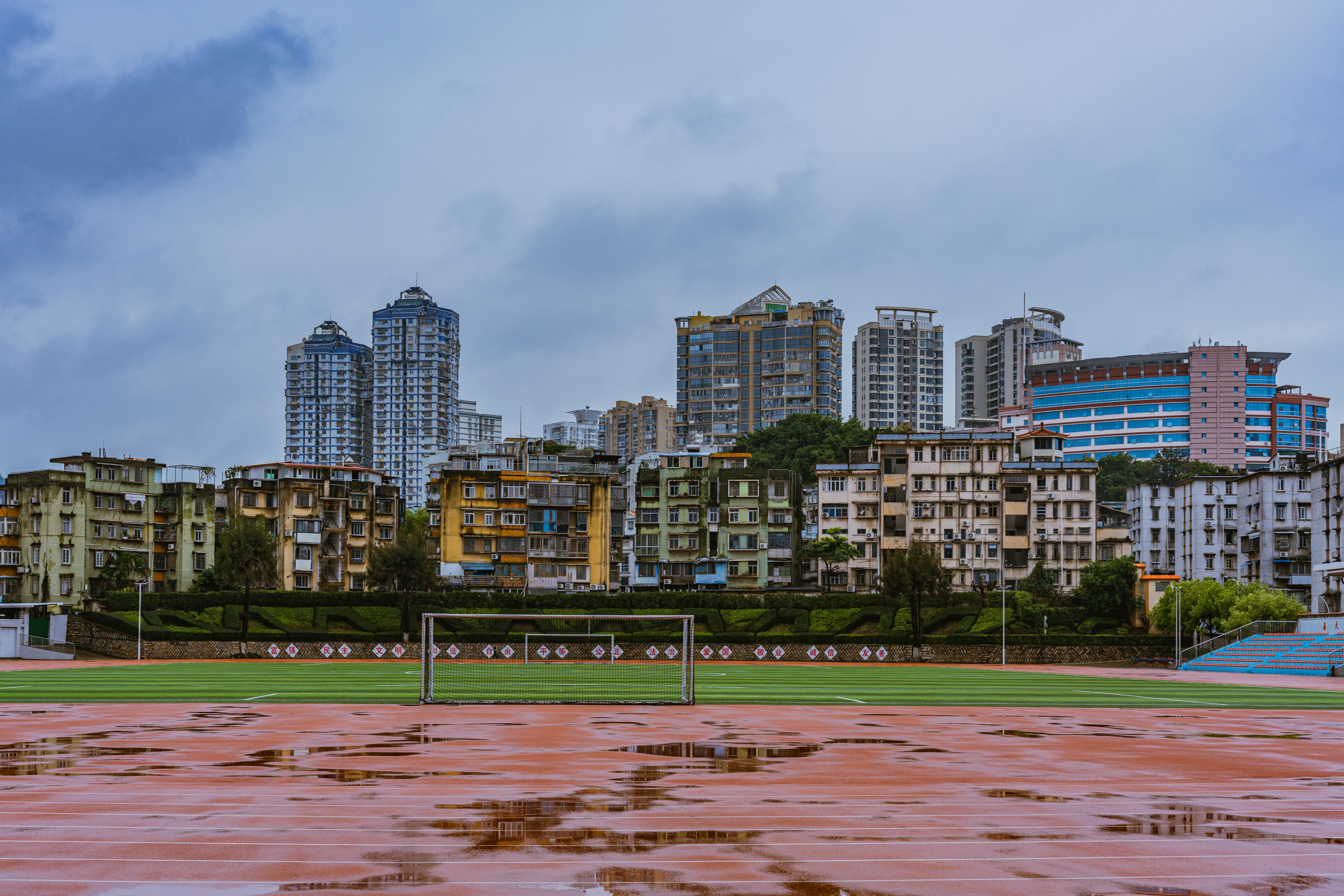 A city skyline with tall buildings behind a football pitch and puddles, captured on a cloudy day.