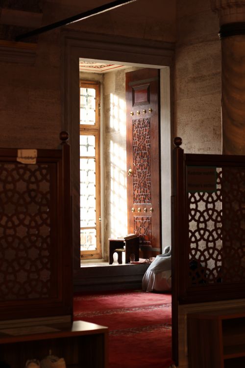 Free Sunlit mosque interior showcasing a traditional carved wooden door and prayer area. Stock Photo