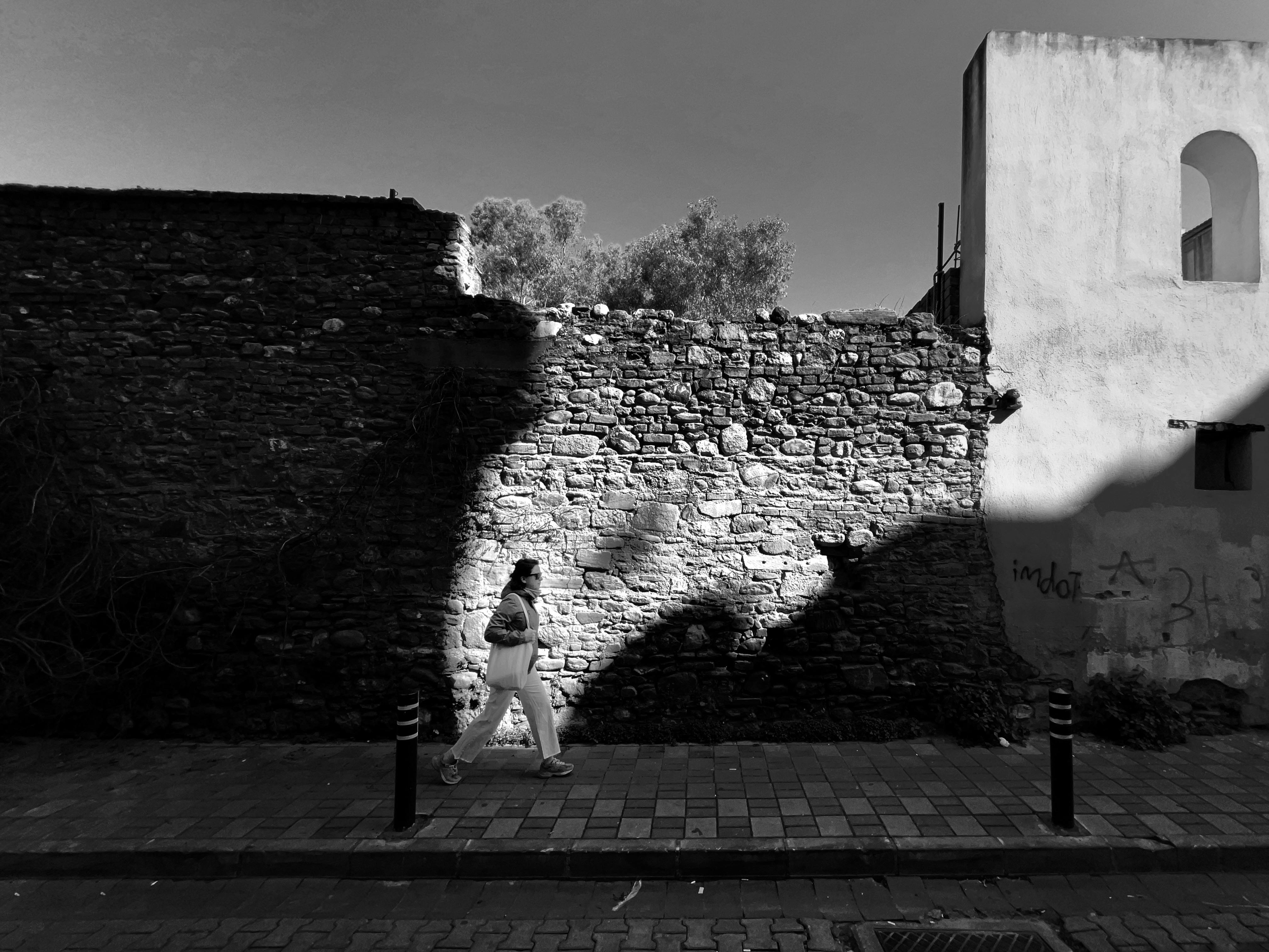 Black and white photo of a woman walking past a historic stone wall in Aydın, Türkiye.