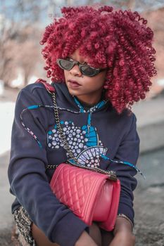 Stylish woman with vibrant red curls and sunglasses, posing outdoors with a quilted handbag.