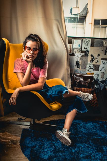 A fashionable young woman in glasses sits thoughtfully in a stylish yellow chair.