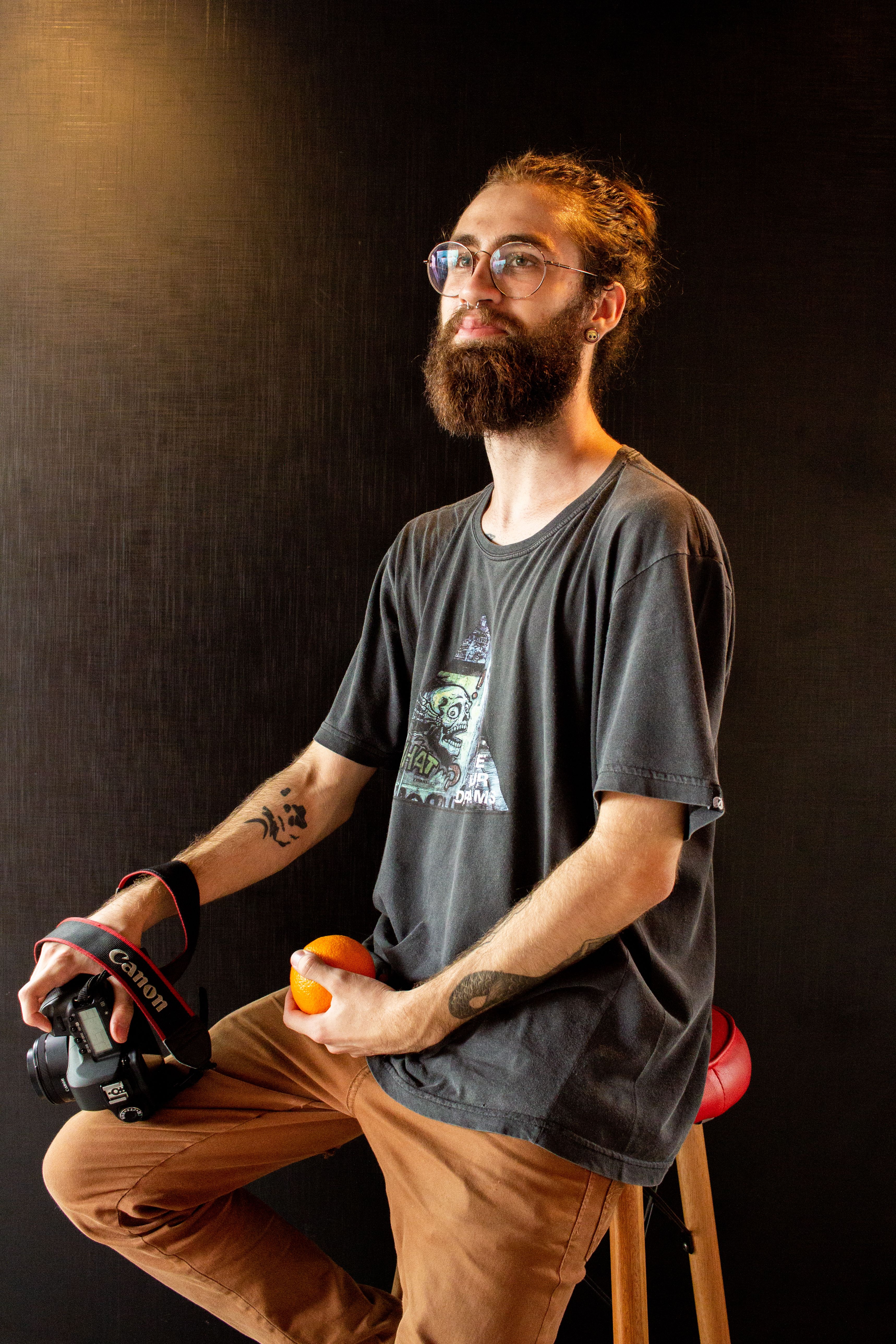 Portrait of a bearded man holding a camera and orange, showcasing a modern hipster style.