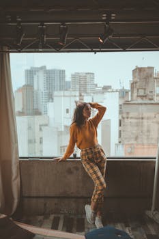 Fashionable woman posing by window with city skyline view, showcasing modern urban style.