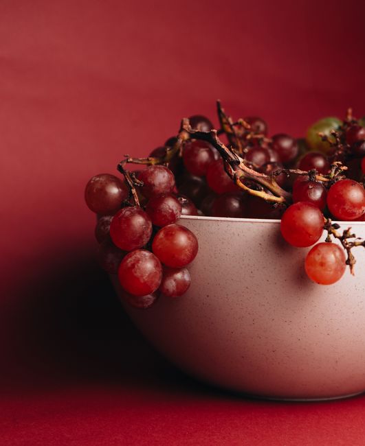 A close-up of juicy red grapes in a ceramic bowl against a rich red background.