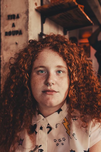 Portrait of a woman with curly red hair and a septum piercing in a stylish cafe setting.