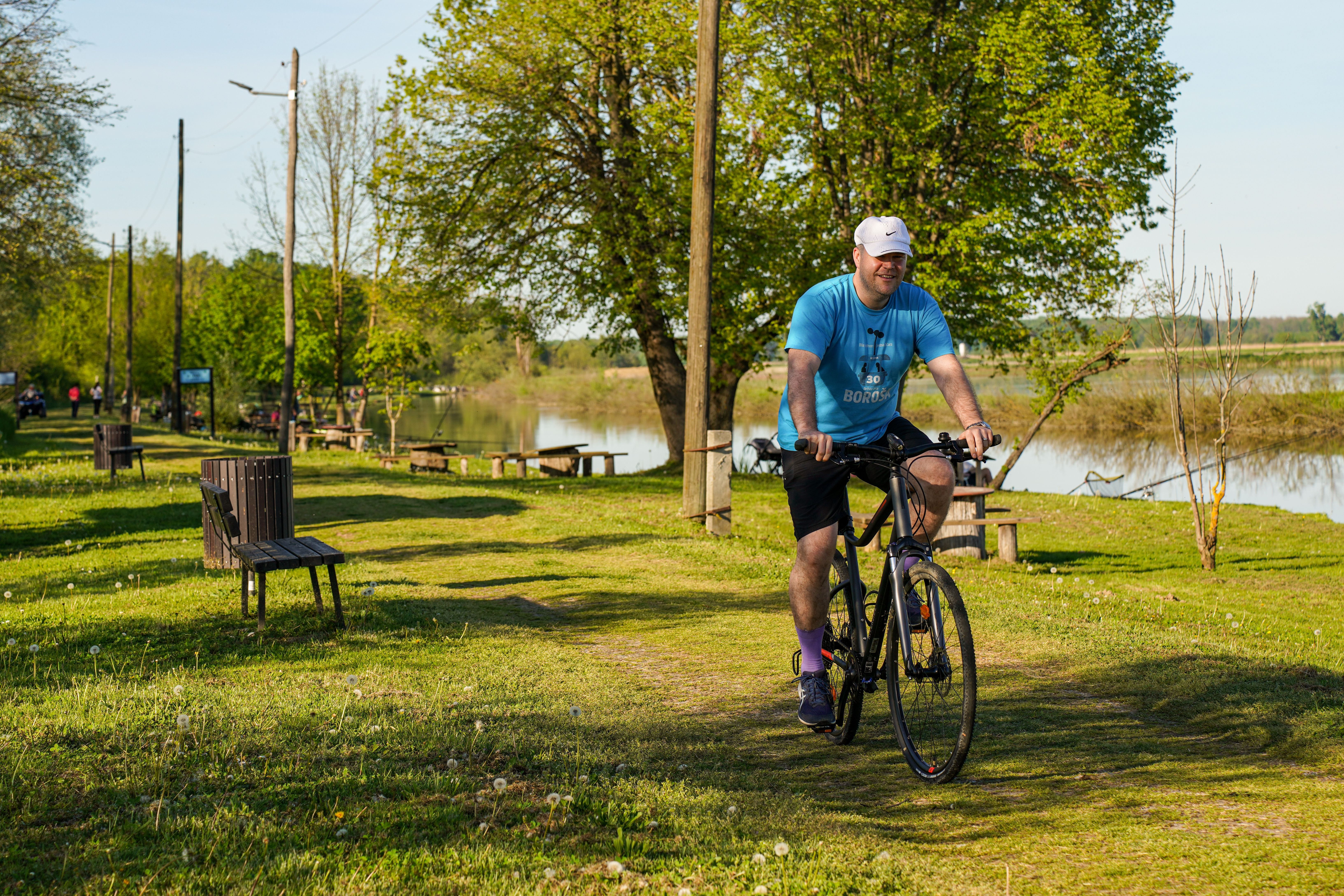 Adult man cycling along a lakeside path in a sunny park setting, surrounded by greenery and benches.