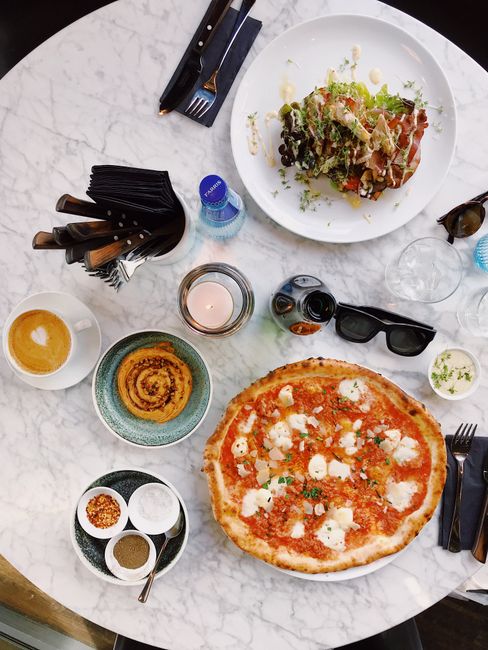 Aerial shot of a dining table with pizza, salad, coffee, and tableware on a marble top.