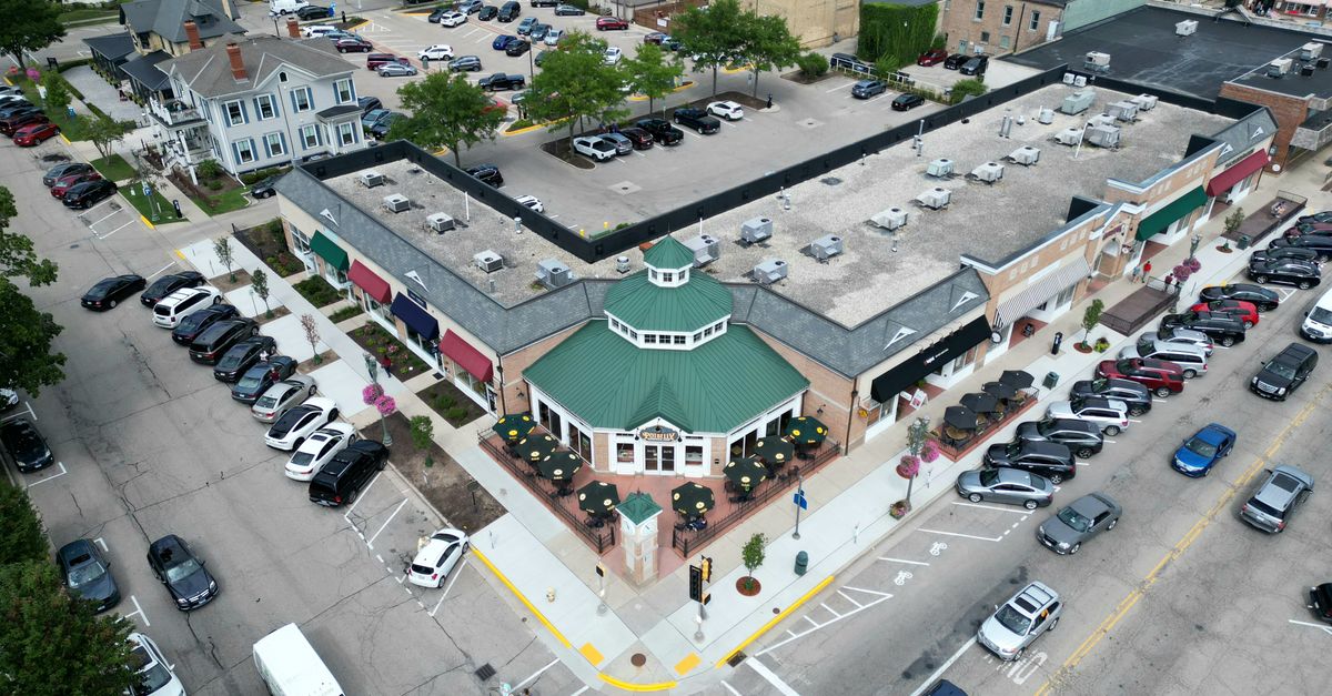 Bird's-eye view of a bustling suburban shopping center with parked cars and tree-lined streets.