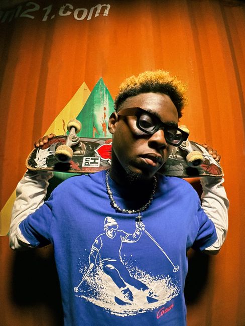 Stylish young man with blonde dyed hair and glasses holding a skateboard against an orange background.