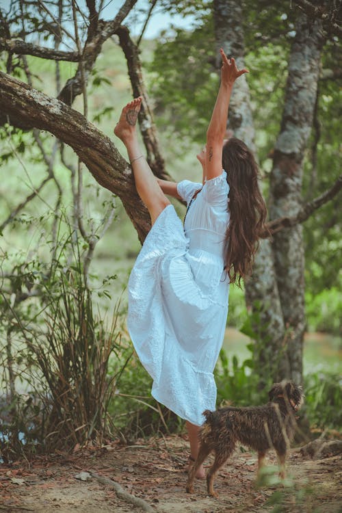 Free Woman Wearing White Dress Puts Her Left Leg on Tree Beside Dog Stock Photo