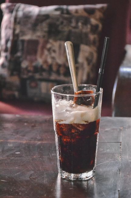 A tall glass of cola with swirls of cream, coconut syrup, and a straw, ice cubes visible through the glass.