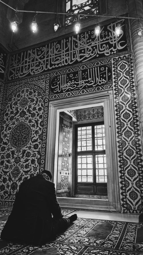Free Black and white photo of a man praying in a beautifully decorated mosque with intricate Arabic calligraphy. Stock Photo