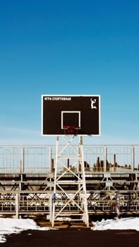 A lone basketball hoop stands outdoors against a wintry urban backdrop under a clear blue sky.