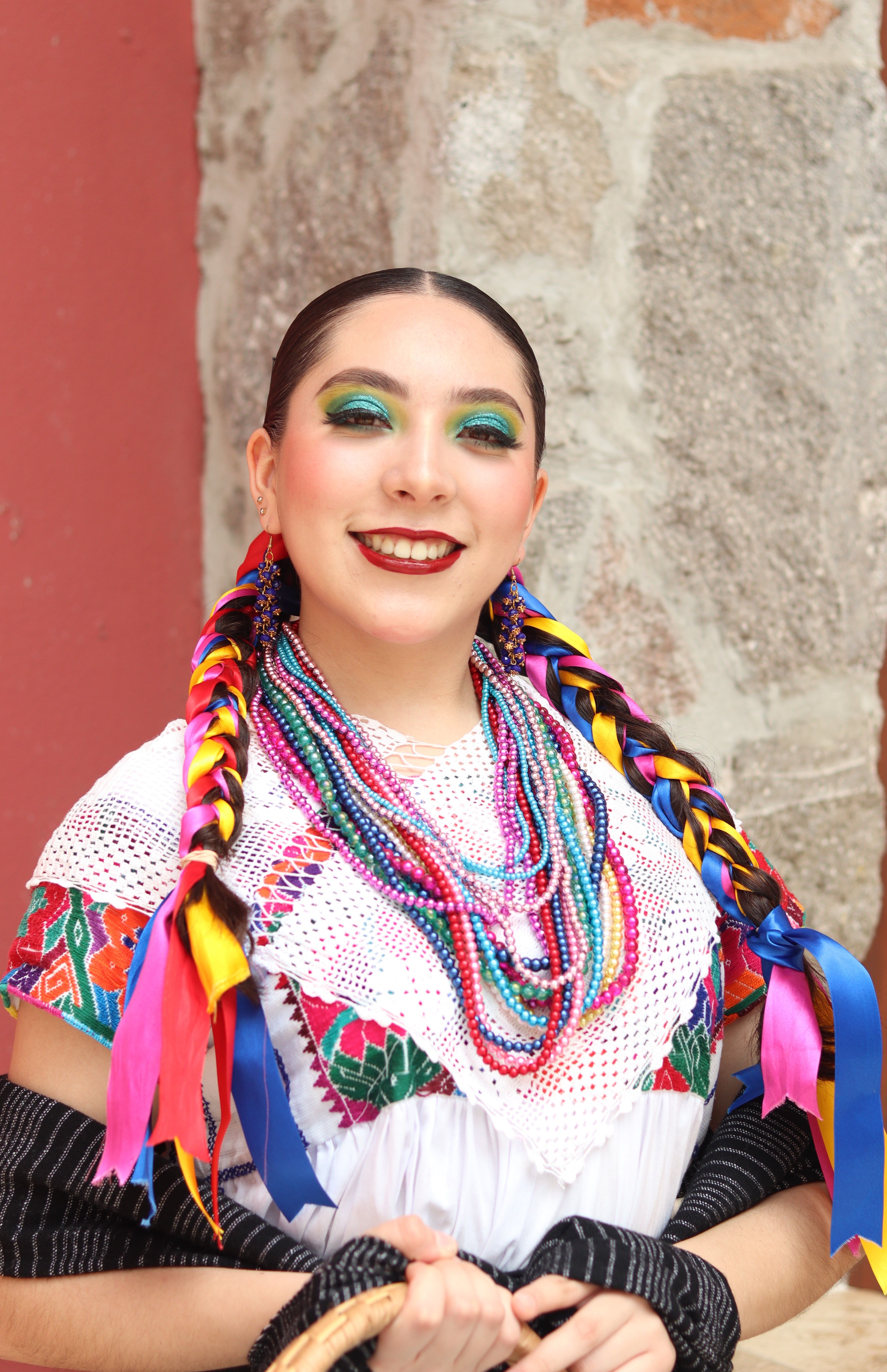 Colorful portrait of a smiling woman in traditional Mexican folk attire with braided ribbons.