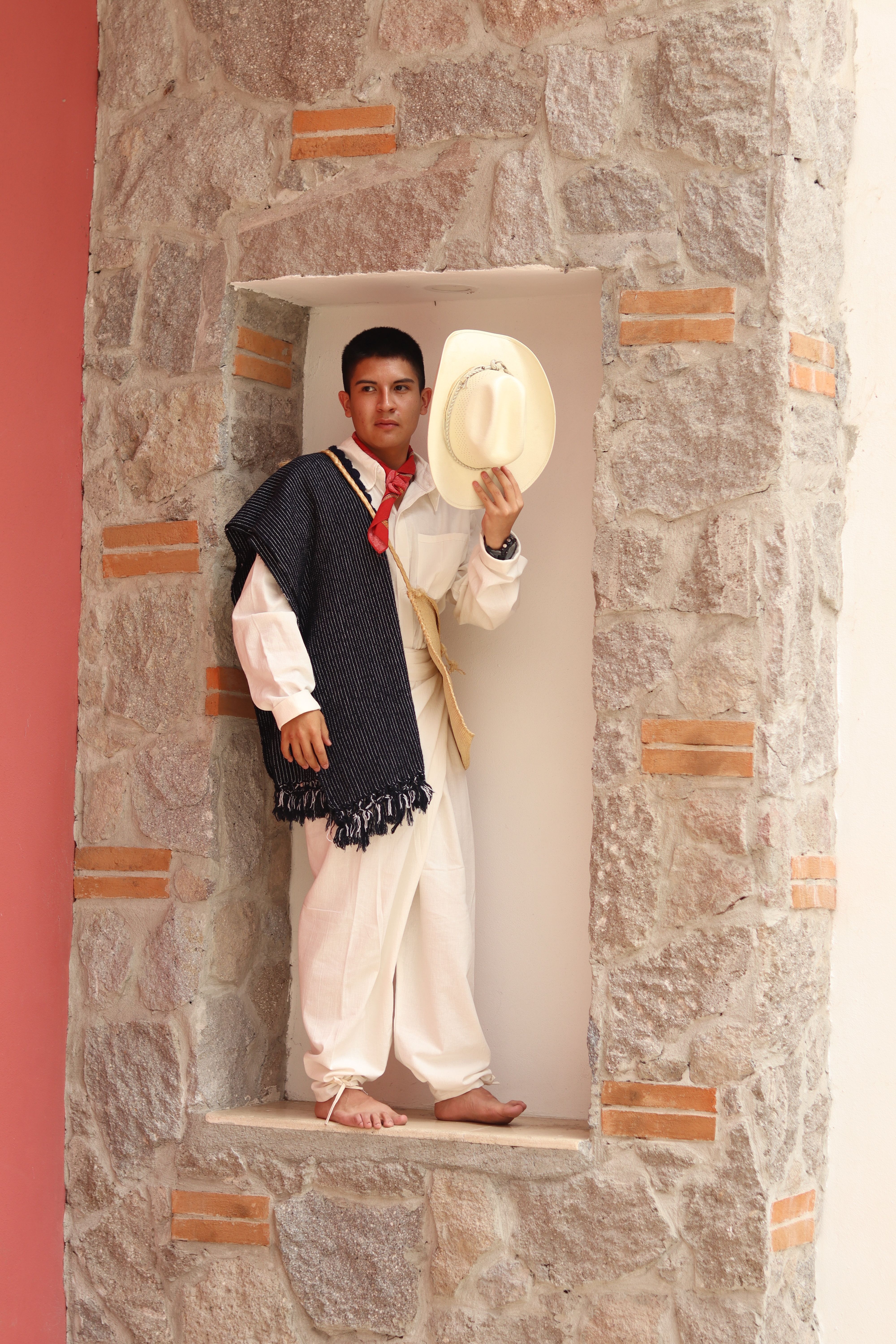 Man in traditional Mexican clothing with sombrero in stone alcove.