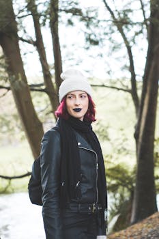 一位粉色头发的时尚女性，身穿皮夹克、头戴无檐帽，置身于树木繁茂的秋景之中