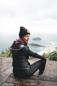 阴天里，一名穿着户外服装的女子从悬崖上欣赏着宁静的海岸景色