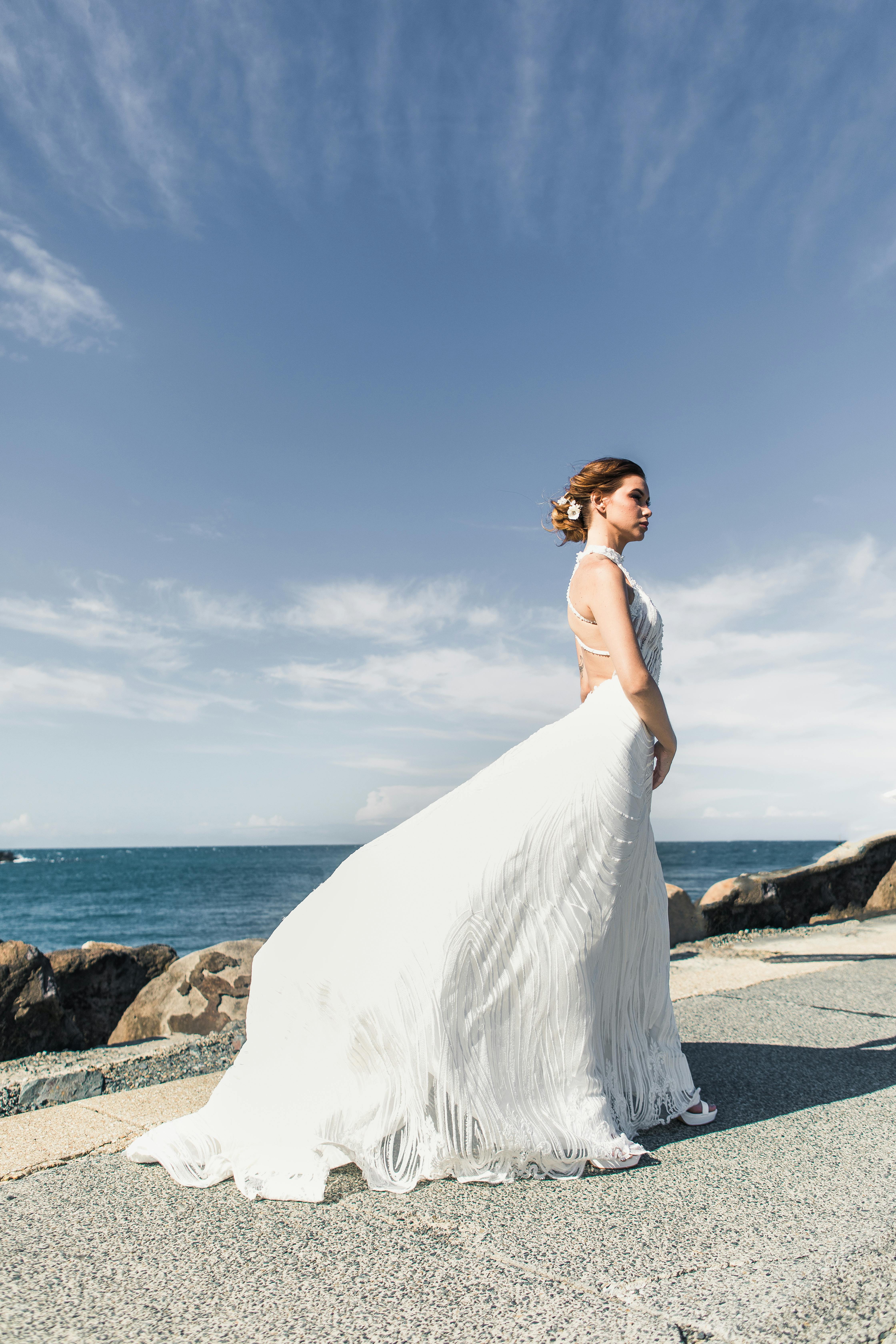 A beautiful bride in a flowing wedding gown poses gracefully by the seaside.