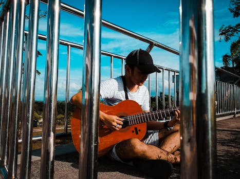 Young man sitting outdoors playing an acoustic guitar, enjoying a sunny day in a park setting.