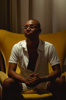 一位戴着眼镜、穿着开领衬衫的年轻人，坐在一把充满活力的黄色椅子上