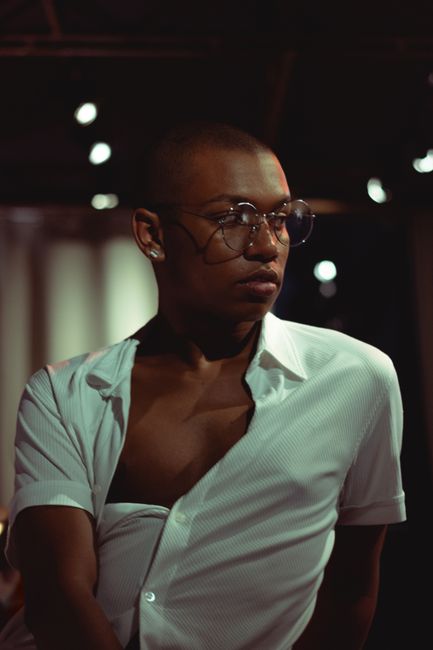 A fashionable man in eyeglasses and open shirt poses confidently indoors.