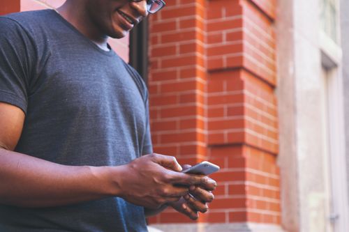 Free A smiling man checks his smartphone while leaning against a brick wall outdoors. Stock Photo