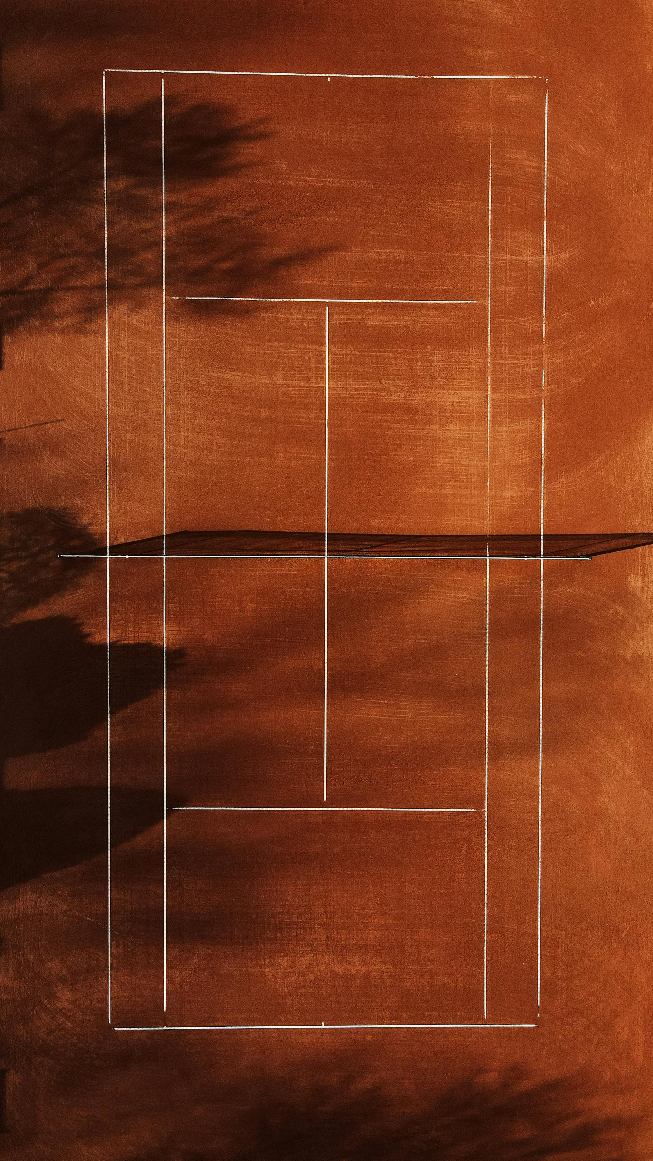 Drone shot of an empty orange clay tennis court with shadows and lines in sunlight.