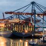 A massive cargo ship loaded with shipping containers is docked at an illuminated port terminal at dusk.