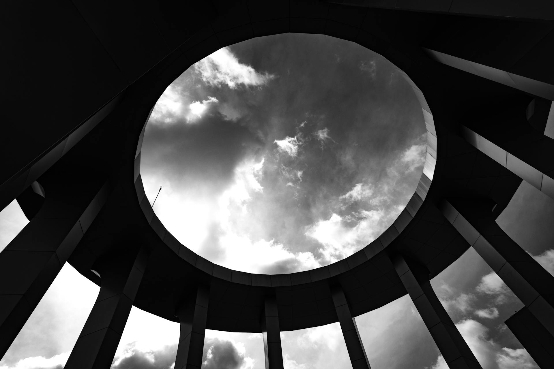 Black and white image of a circular architectural skylight framing a cloudy sky.