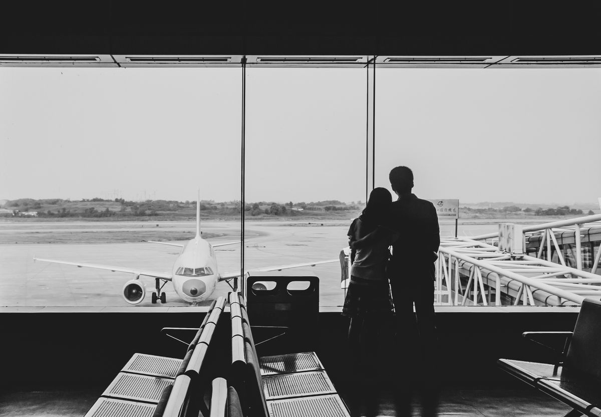 Silueta unui cuplu imbratisat la geamul unui terminal, cu avion pe pista