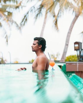 Man enjoying an orange cocktail at an infinity pool under palm trees in Maldives resort.