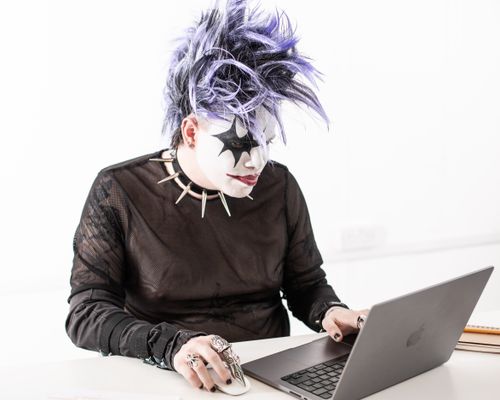 Free A person with costume makeup and purple hair working on a laptop against a white background. Stock Photo