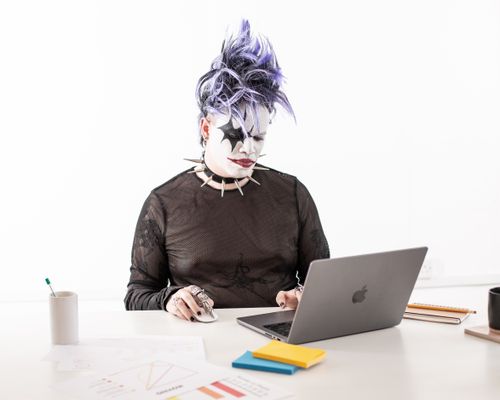 Free A goth-styled man with purple hair works on a laptop in a bright studio setting, showcasing modern Halloween office vibes. Stock Photo