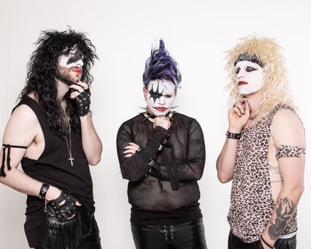 Three adults in glam rock attire and makeup pose on a white background.
