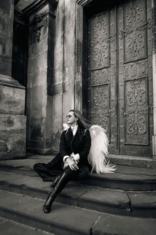 Free Graceful woman in angel costume sitting on ancient steps in Lviv. Black and white artistry enhances the serene atmosphere. Stock Photo