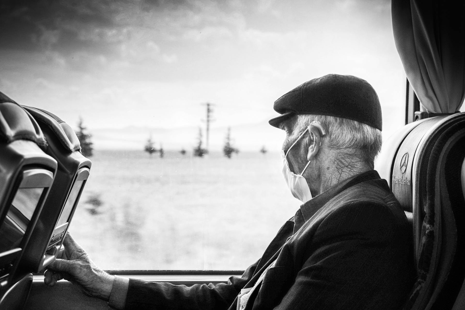 Elderly man wearing a mask, gazing out a bus window, capturing a serene travel moment.