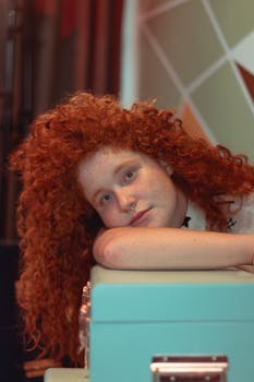 这幅迷人的肖像描绘了一位留着红色卷发的女人，她正靠在手提箱上