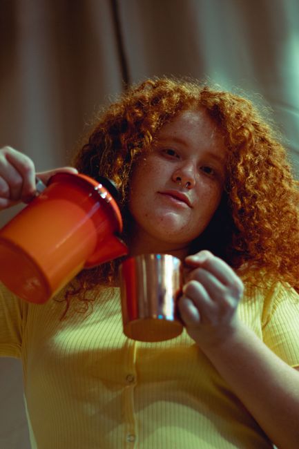A young woman with curly red hair pouring tea from a teapot into a cup. Indoor setting.