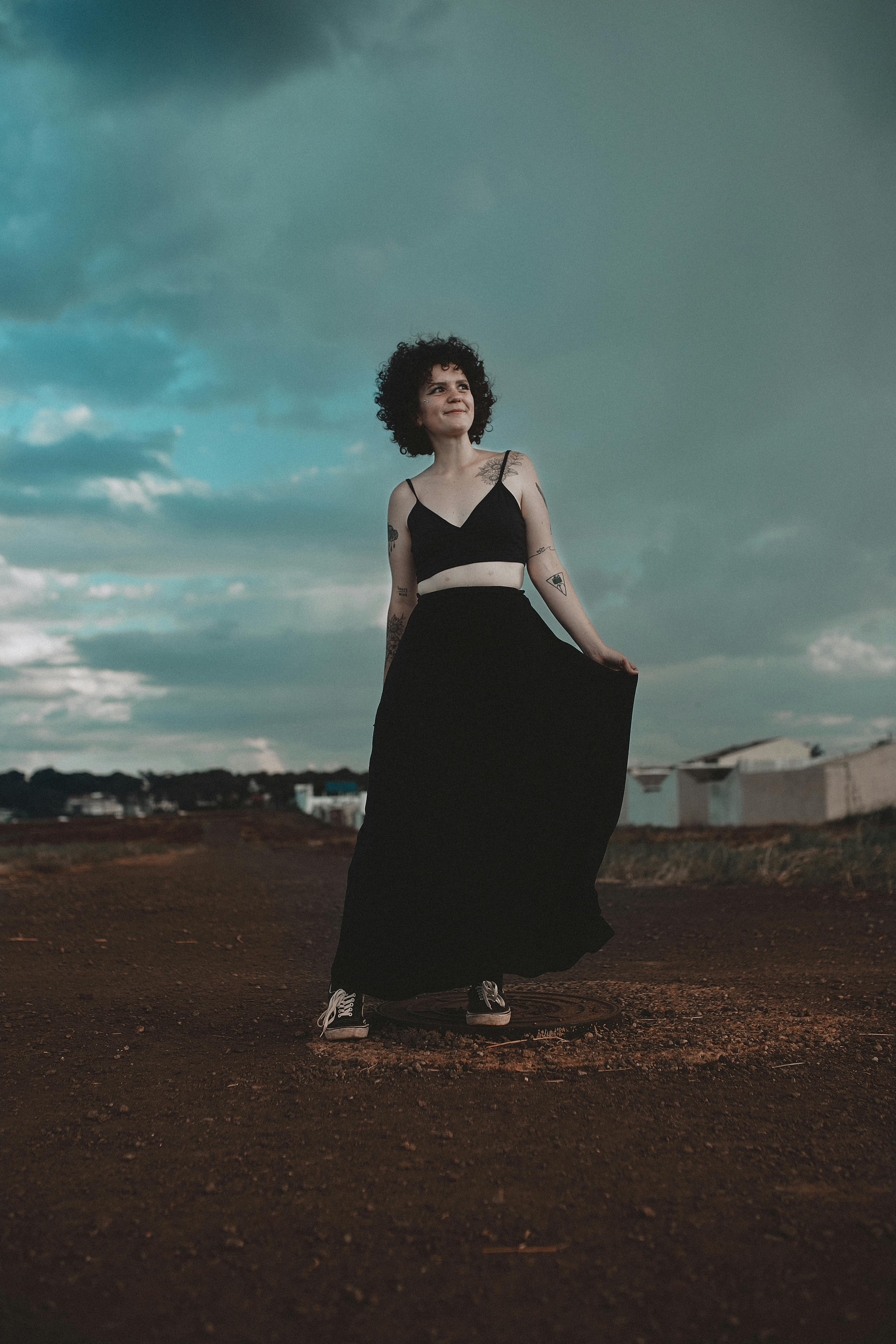 一名卷发纹身的女子身着黑色连衣裙，在戏剧性的天空下于户外摆姿