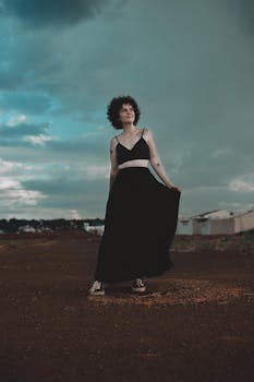 一名卷发纹身的女子身着黑色连衣裙，在戏剧性的天空下于户外摆姿